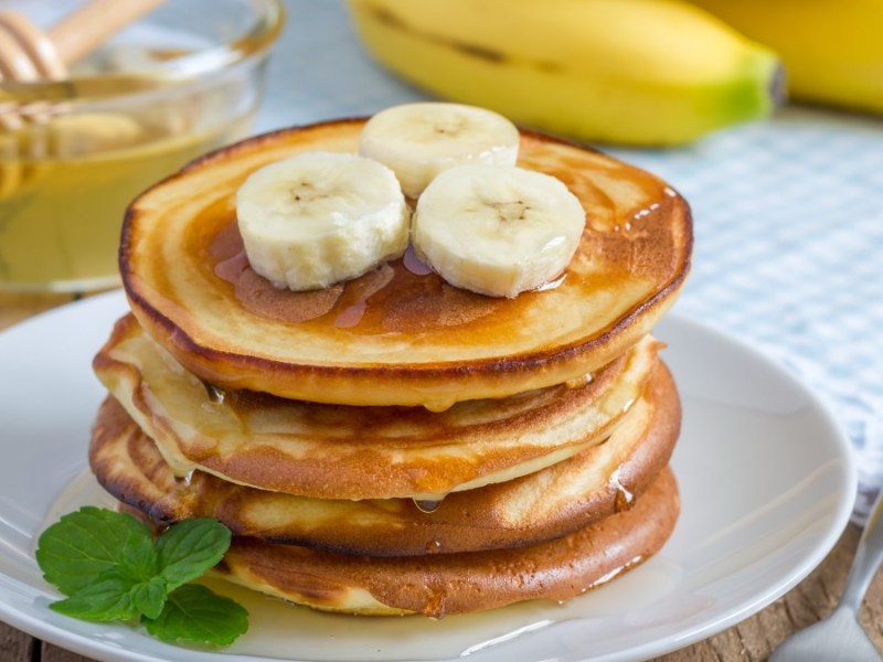 Ein Stapel Bananen-Pancakes mit Ahornsirup und Bananenscheiben. Im Hintergrund liegen Bananen.