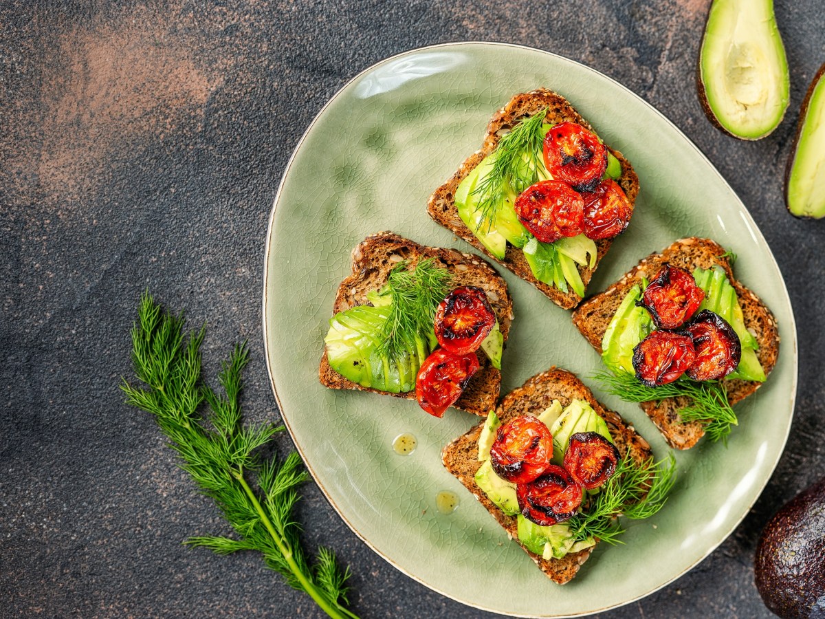 3 Avocado-Brote mit Ofentomaten und KrÃ¤utern belegt auf einem Teller in der Draufsicht, drumherum ganze und halbierte Avocados.