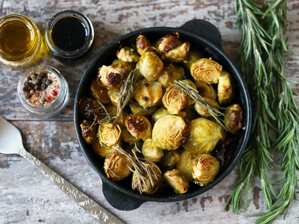 Gebackener Rosenkohl mit Balsamico und Ahornsirup in einer gusseisernen Form in der Draufsicht, daneben GlÃ¤ser mit Zutaten und Rosmarin.