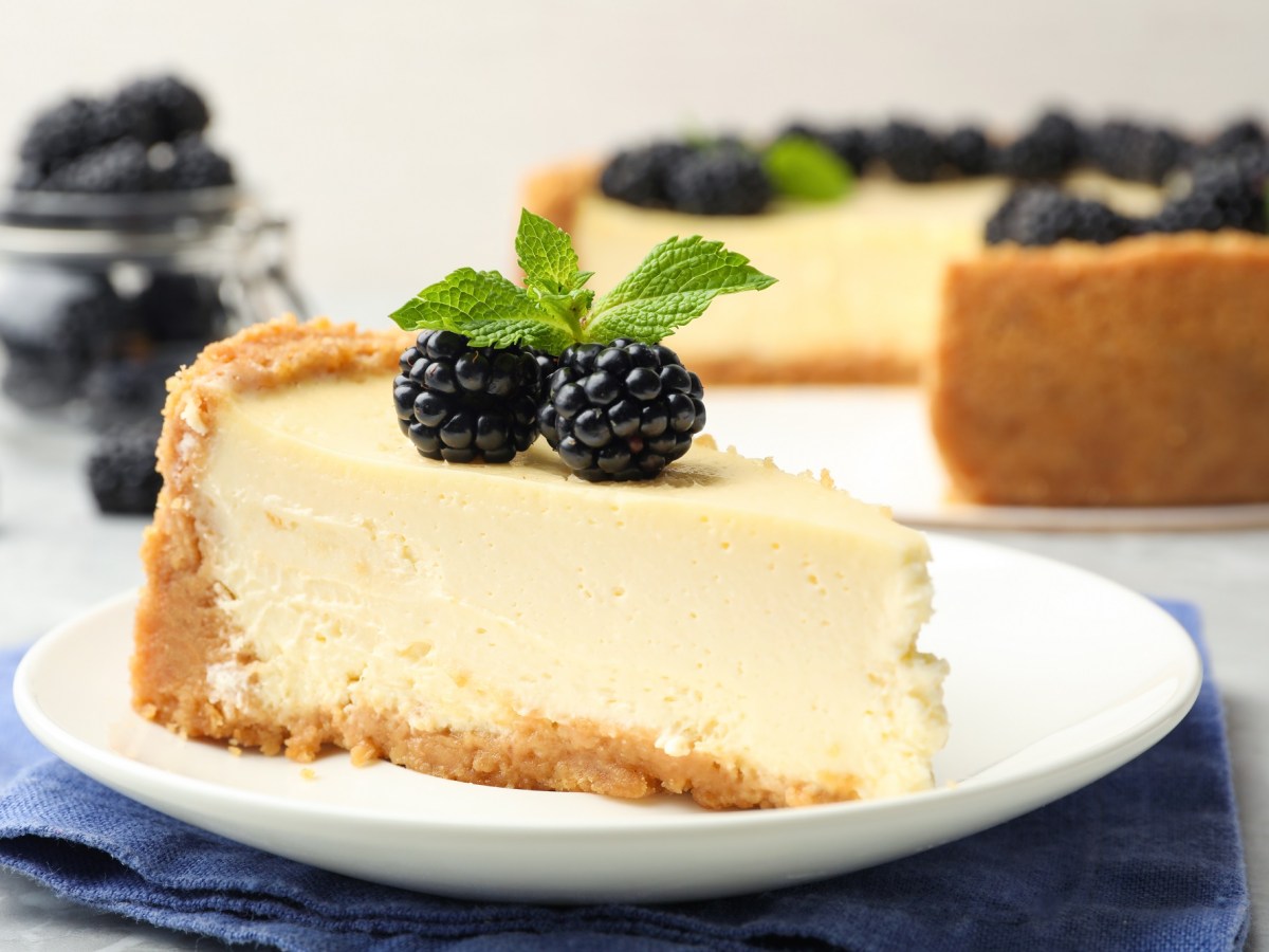 Ein Stück veganer Käsekuchen auf einem Teller, garniert mit Minze und Brombeeren, dahinter unscharf der angeschnittene Kuchen.