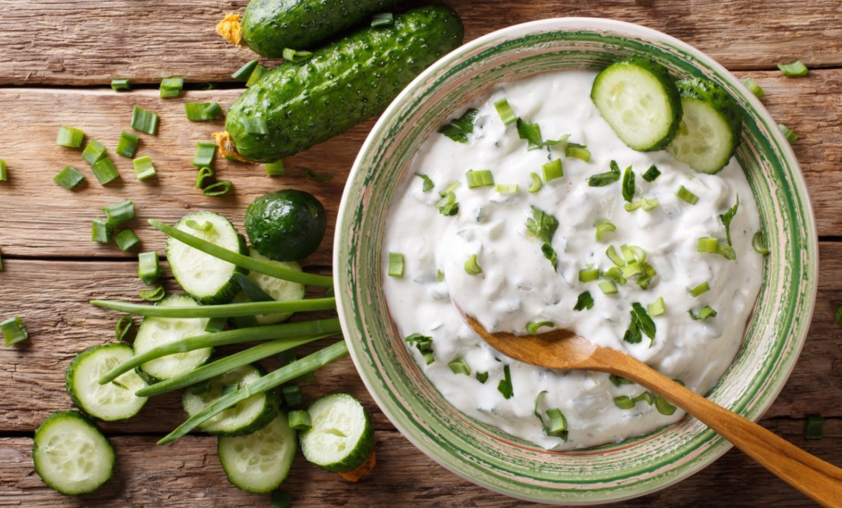 Eine Schüssel mit Gurken-Raita, in dem ein Holzlöffel liegt. Daneben liegen Gurken und frische Zutaten.