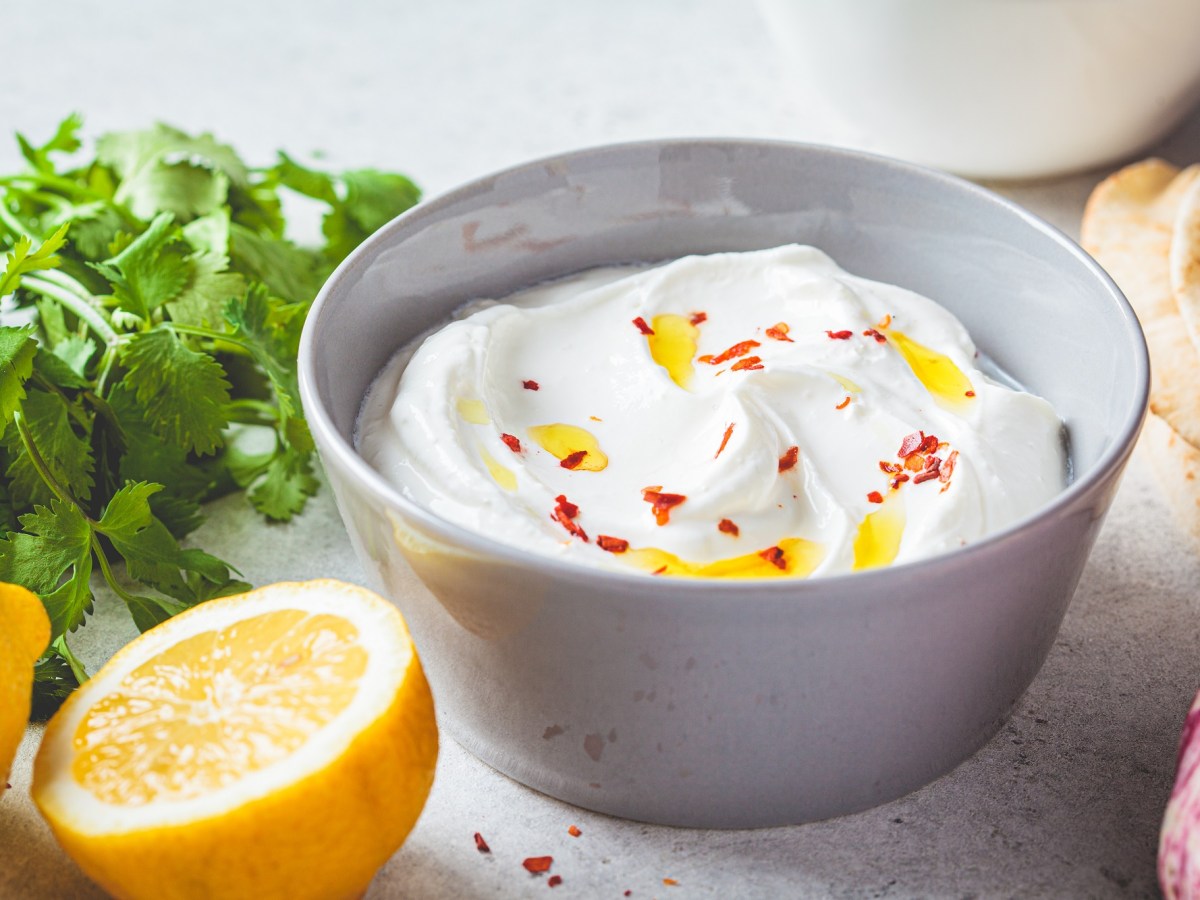 Eine SchÃ¼ssel Feta-Knoblauch-Creme mit Ã–l und Chiliflocken garniert, daneben frische KrÃ¤uter und eine halbierte Zitrone sowie Fladenbrot.