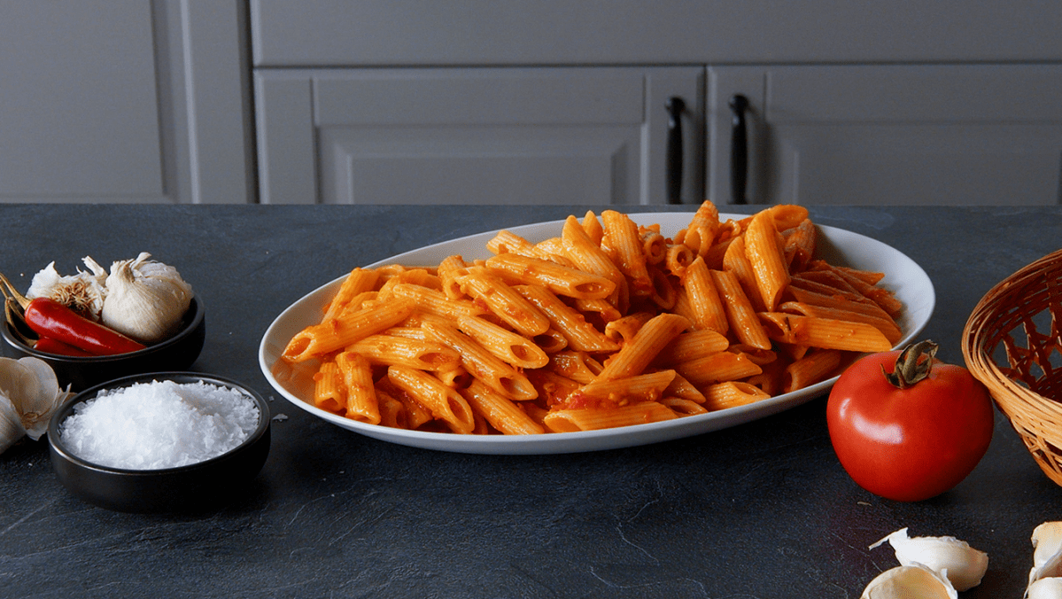 Penne Arrabiata, angerichtet auf einem Teller. Tomaten und Gewürze liegen außen herum.