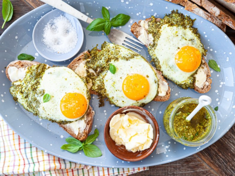 Drei Pesto-Eier auf Brotscheiben. Drumherum stehen Schalen mit Pesto, Butter und Salz.