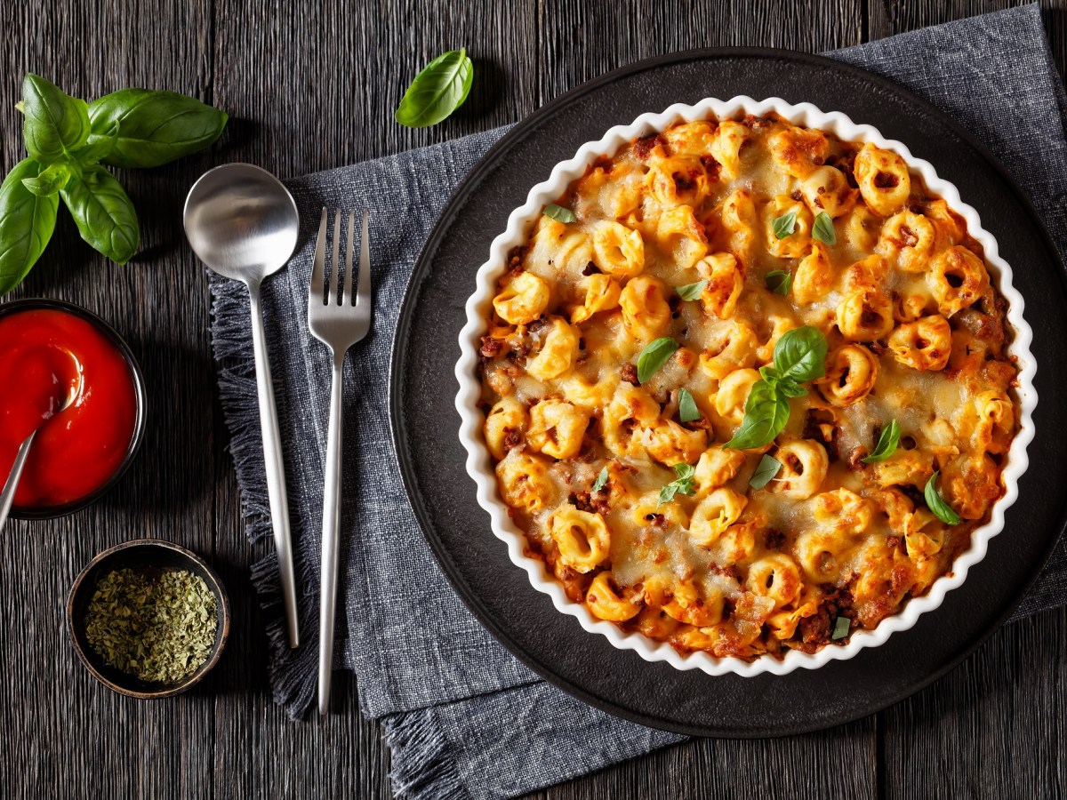 Tortellini-Auflauf in einer geriffelten Auflaufform auf einem schwarzen Tablett. Darunter ein graues Platzdeckchen, Löffel und Gabel und Tomatensoße. Draufsicht.