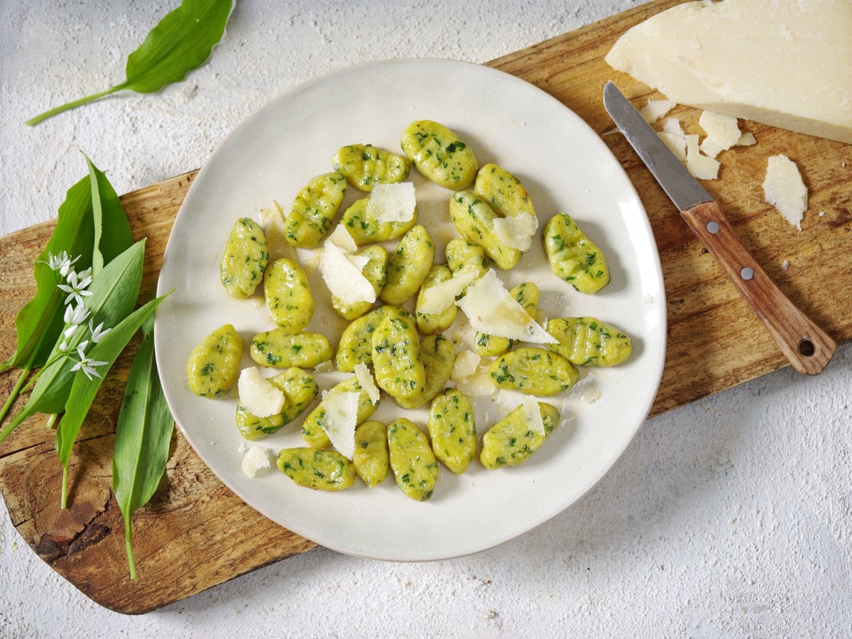 Ein Teller Bärlauch-Gnocchi, die mit Parmesan bestreut sind, auf einem Brett, daneben ein Stück Käse und Bärlauchblätter. Draufsicht.