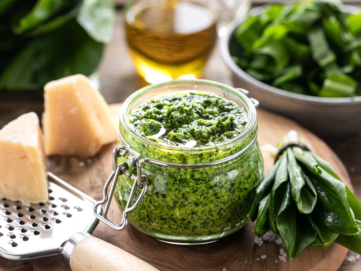 ein Glas Bärlauchpesto, daneben frischer Bärlauch und Parmesan sowie eine Käsereibe und eine Olflasche.