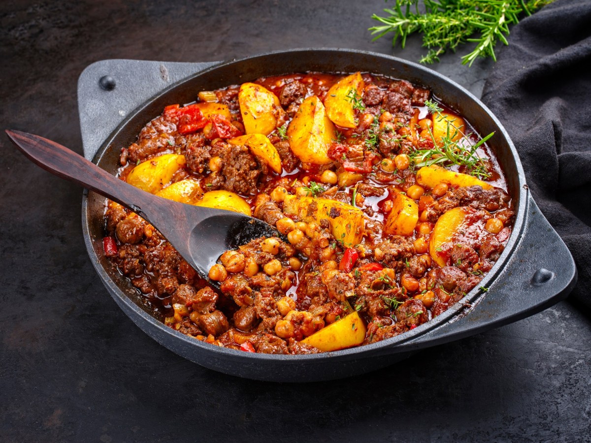 Bauerntopf mit Hackfleisch, Kartoffeln, Paprika und mehr Gemüse in einer tiefen Pfanne, darin ein Holzlöffel, Nahaufnahme.