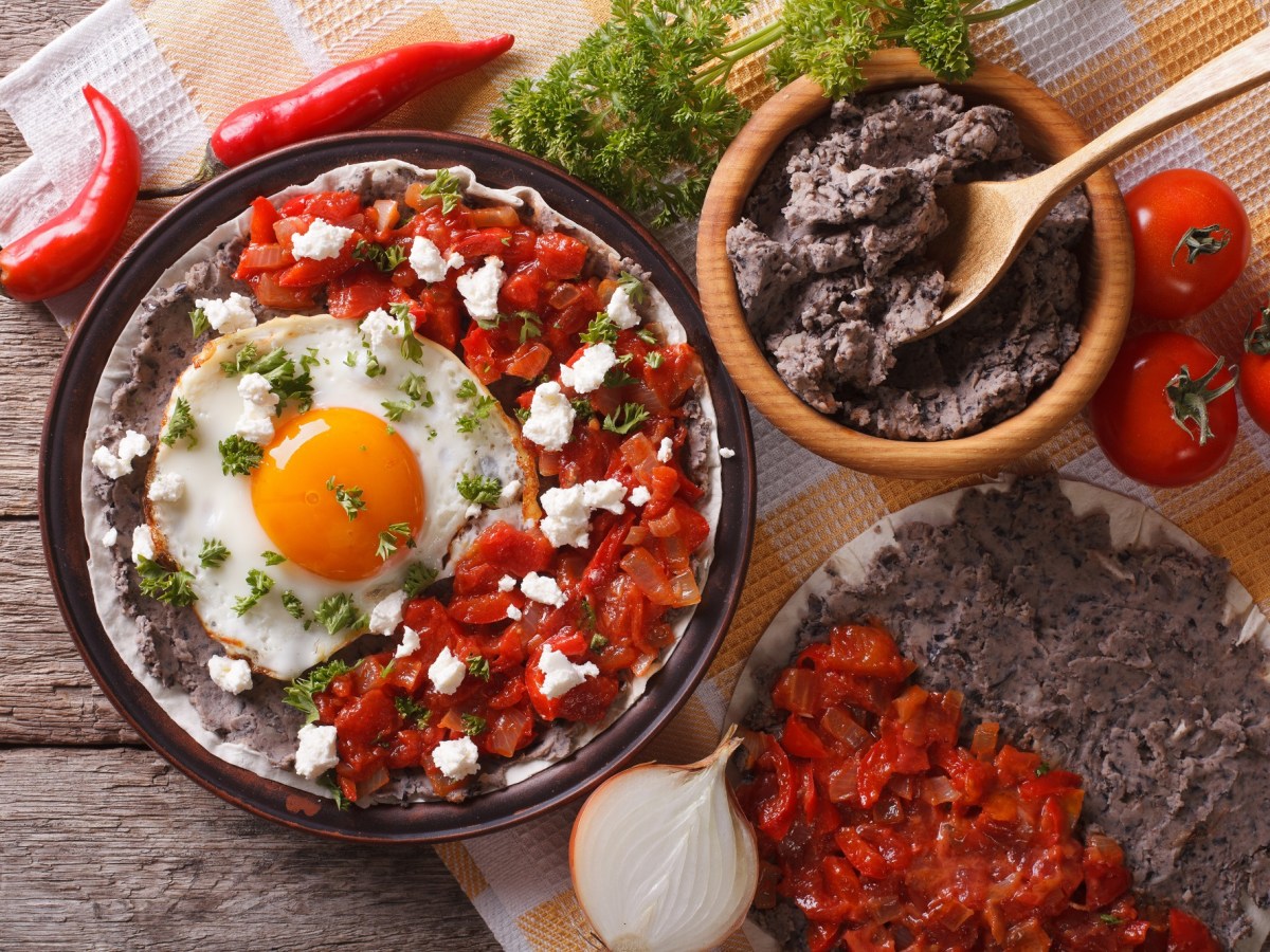 Huevos Rancheros auf einem dunklen Teller. Daneben eine Schale mit Bohnenmus, eine weitere Tortilla mit Tomatensoße und Bohnenmus und Tomaten, Chilis, Zwiebeln und Petersilie. Draufsicht.