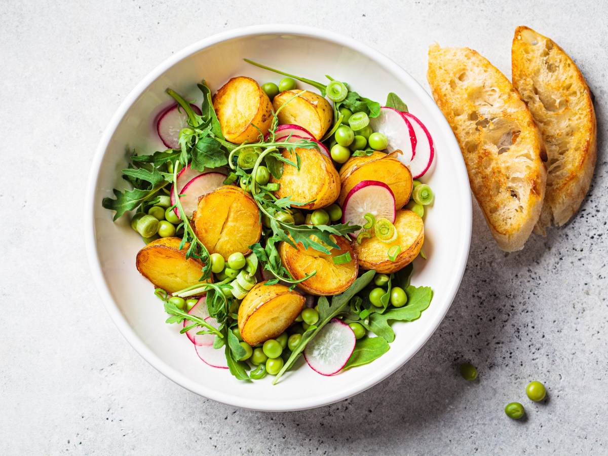 Kartoffelsalat mit Erbsen, Radieschen und Rucola in einer weißen Schüssel. Daneben geröstetes Brot.