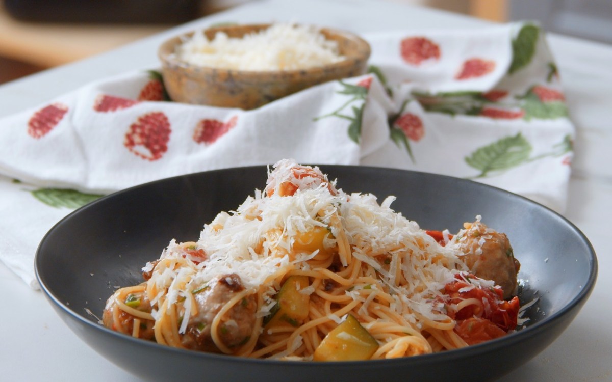 Ein schwarzer Teller mit einer Pasta-Pfanne mit Fleischbällchen, Gemüse und geriebenem Parmesan.