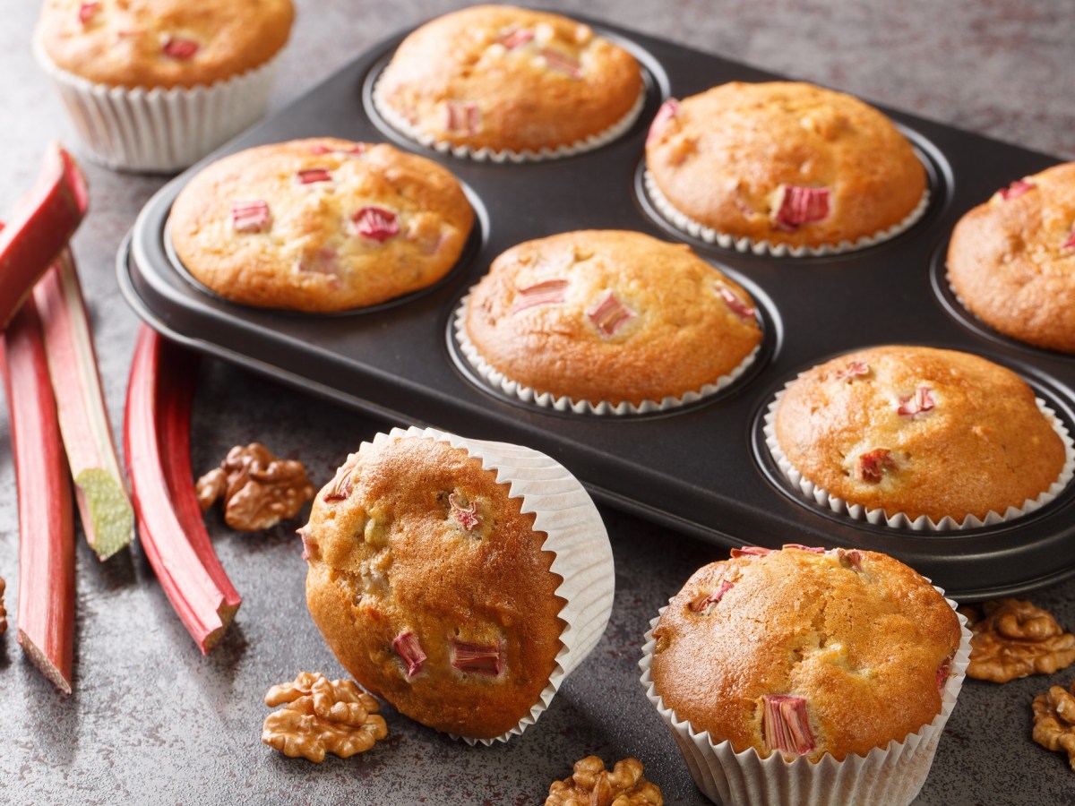 Ein Muffinblech mit Rhabarber-Walnuss-Muffins, zwei Muffins liegen daneben, außerdem Rhabarberstangen und Walnüsse daneben.