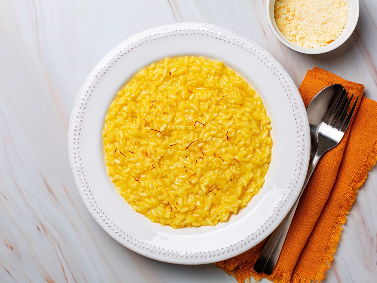 Risotto alla Milanese in einem weißen Teller. Daneben liegen Gabel und Löffel sowie eine orange Serviette. Eine Schale mit Parmesankäse steht auch dabei.