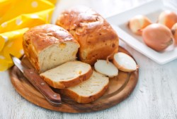 Zwiebelbrot: Zwei Laibe auf einem Holzbrett, eines davon ist angeschnitten. Im Hintergrund liegen Zwiebeln