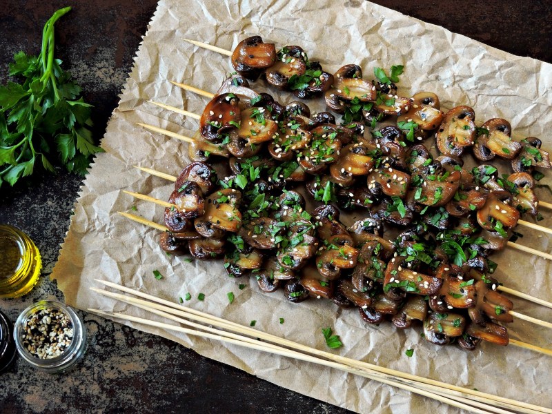 Champignon-Grillspieße auf einem Backpapier, bestreut mit Sesam und Petersilie.