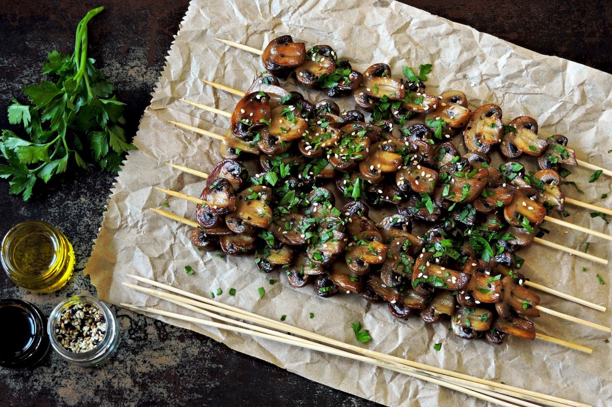 Champignon-Grillspieße auf einem Backpapier, bestreut mit Sesam und Petersilie.