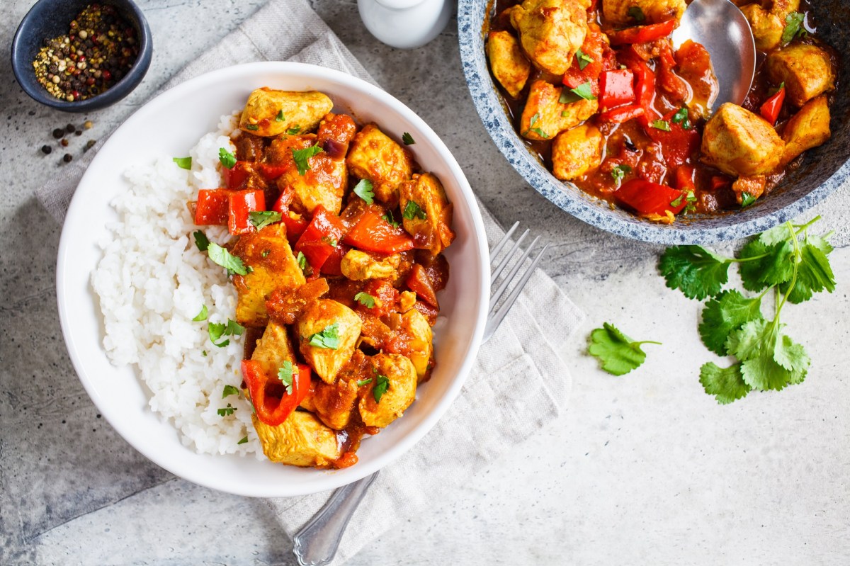 Eine Portion Chicken Jalfrezi mit Basmiatireis in einer weißen Schüssel. Daneben eine Pfanne mit mehr von dem gericht.