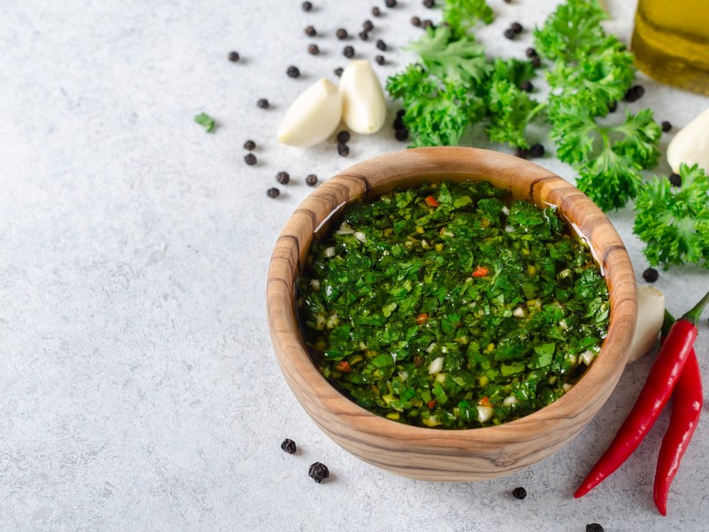 Eine Holzschale Chimichurri, daneben Knoblauchzehen, Chilischoten, Kräuter und Pfefferkörner.