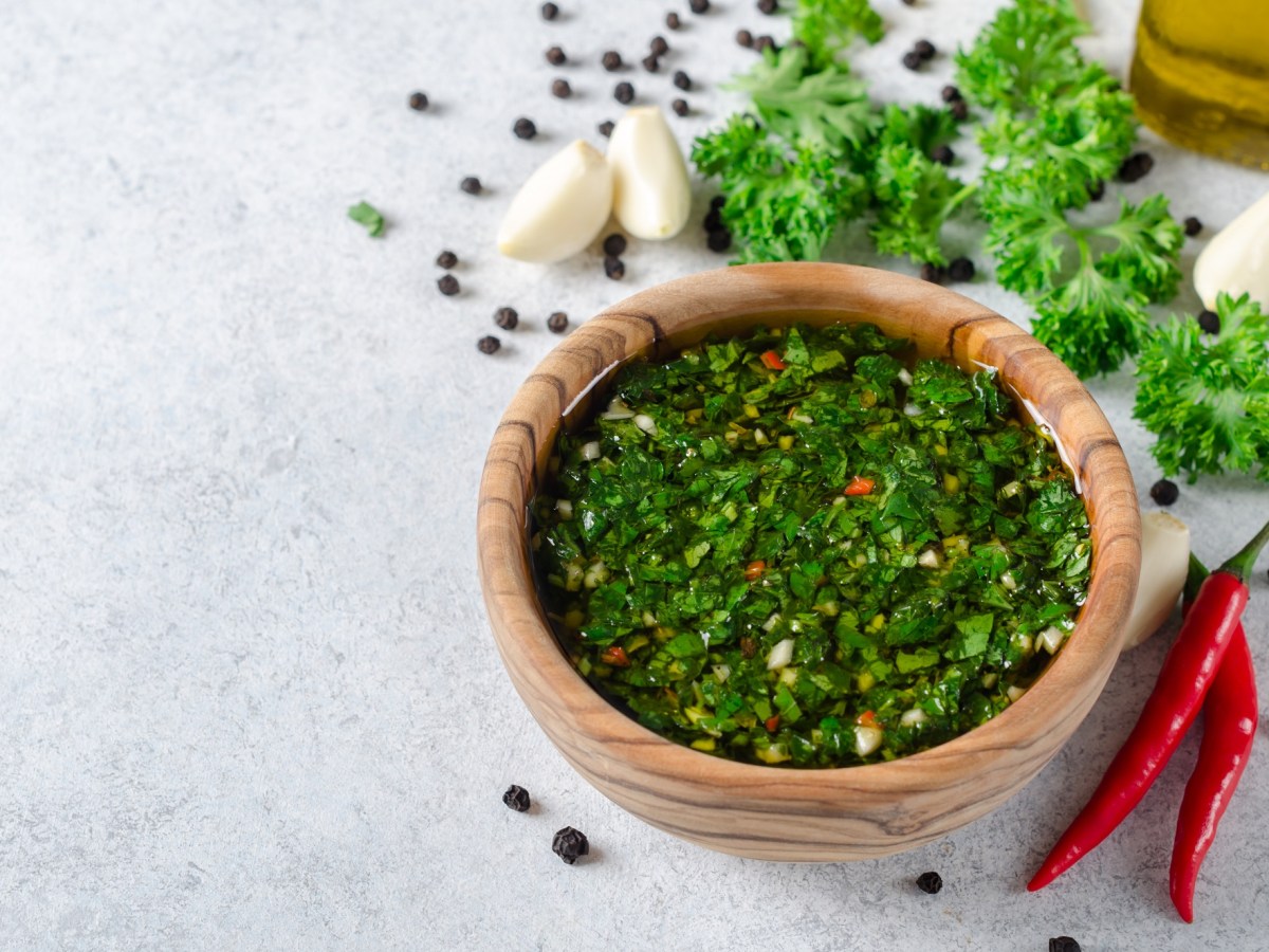 Eine Holzschale Chimichurri, daneben Knoblauchzehen, Chilischoten, Kräuter und Pfefferkörner.