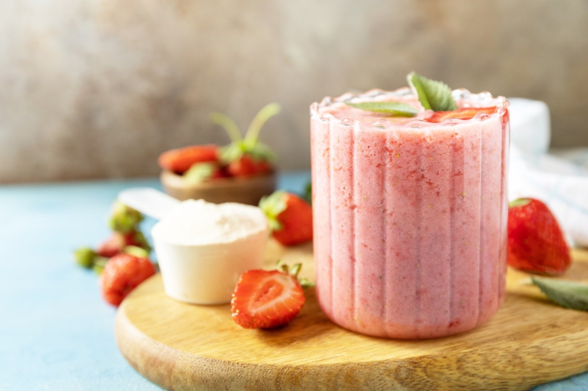 Ein Glas Erdbeer-Sahne-Shake mit Minze garniert auf einem Brett, daneben frische Beeren und Sahne in einer Schüssel.