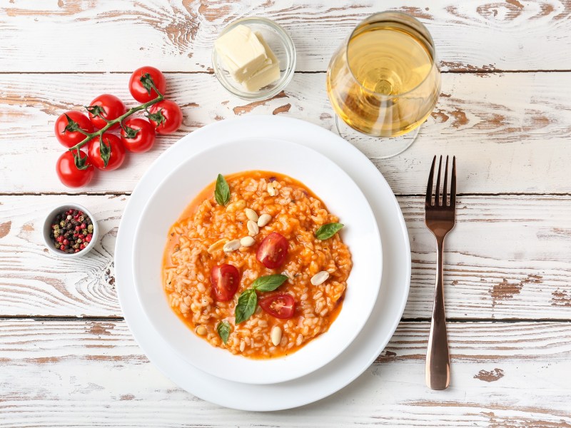 Graupenrisotto mit Tomaten auf einem weißen, tiefen Teller. Daneben stehen ein Glas Weißwein ,einige Kirschtomaten und etwas Butter.
