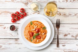 Graupenrisotto mit Tomaten auf einem weißen, tiefen Teller. Daneben stehen ein Glas Weißwein ,einige Kirschtomaten und etwas Butter.