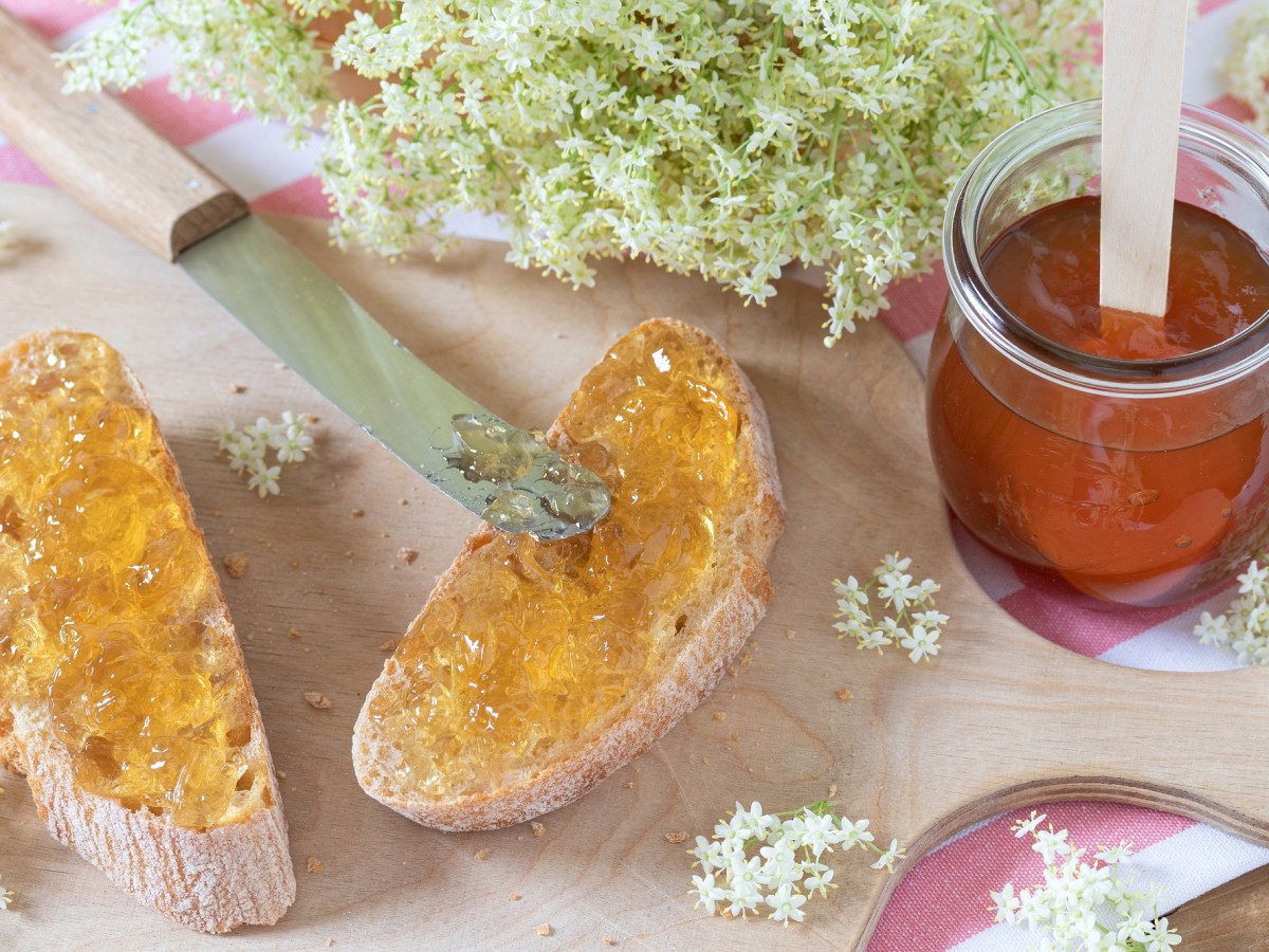 Zwei Brote mit HolunderblÃ¼ten-Gelee, ein Glas Gelee und HolunderblÃ¼ten daneben.