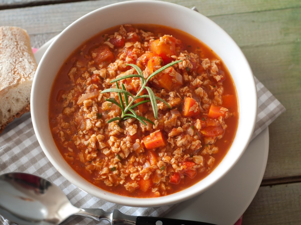 Käse-Hack-Suppe mit Tomaten und Möhren in einer Schüssel, daneben eine karierte Serviette, ein Löffel und ein Stück Baguette.