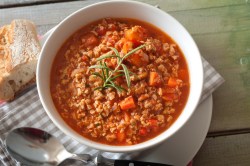 Käse-Hack-Suppe mit Tomaten und Möhren in einer Schüssel, daneben eine karierte Serviette, ein Löffel und ein Stück Baguette.