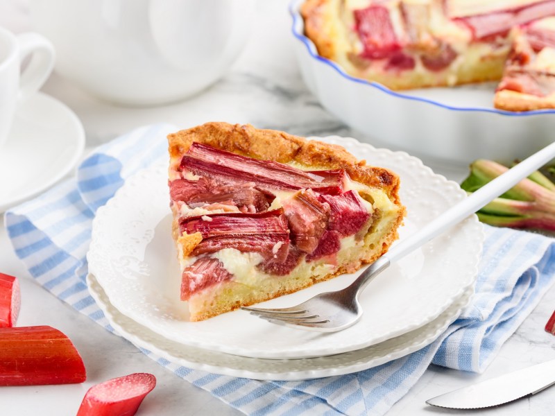 Ein Stück Rhabarber-Pudding-Tarte auf einem Teller mit Kuchengabel, dahinter die Kuchenform.