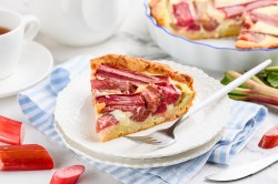 Ein Stück Rhabarber-Pudding-Tarte auf einem Teller mit Kuchengabel, dahinter die Kuchenform.