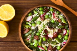 Eine Holzschüssel (Öffnet in einem neuen Browser Tab) Spargel-Couscous-Salat mit Erbsen und Radieschen in der Draufsicht, daneben eine halbierte Zitrone und ein Holzlöffel.