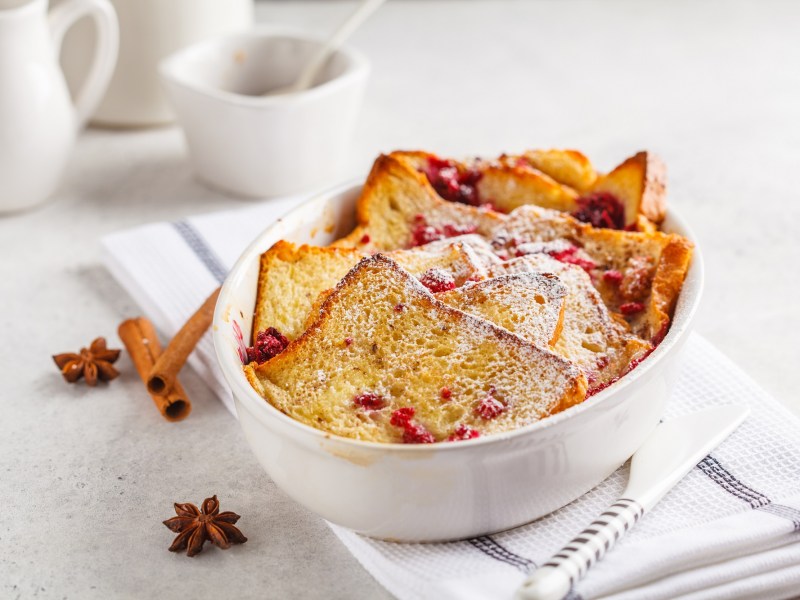 Toastbrotauflauf mit Himbeeren und Puderzucker in weißer Auflaufform, daneben Anissterne und Zimtstangen.