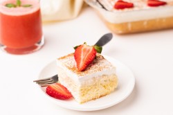 ein Stück Tres Leches Kuchen mit Erdbeeren auf einem weißen Teller. Dahinter steht unscharf der Rest des Kuchens