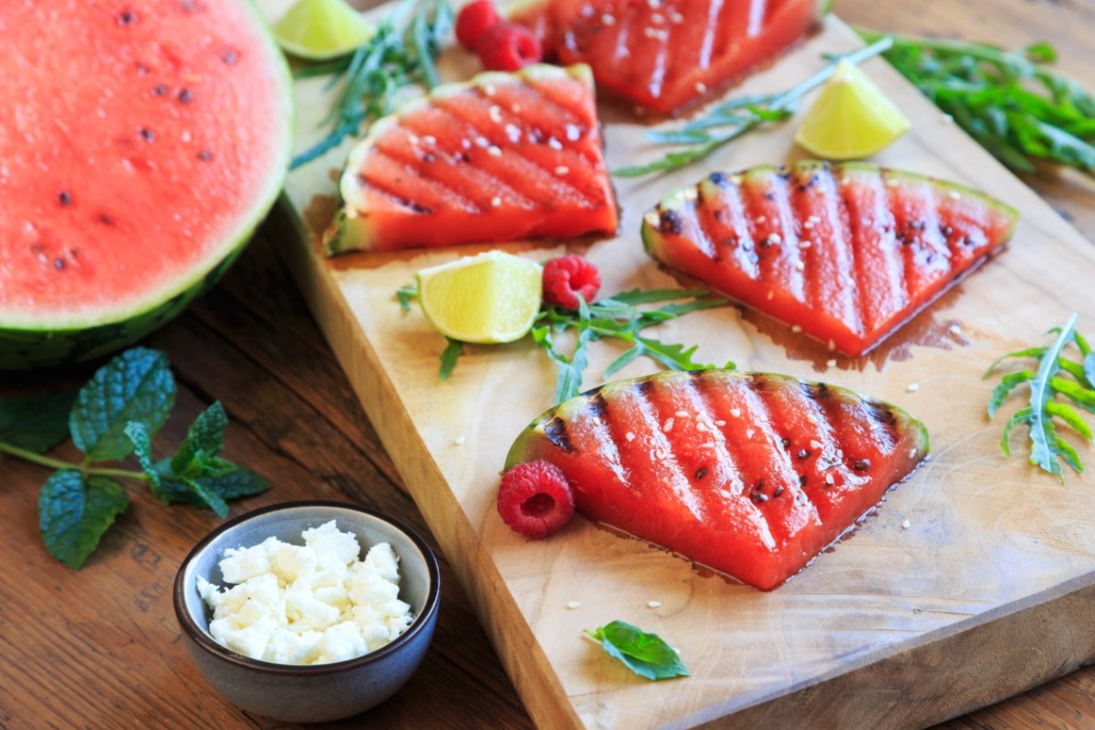 Wassermelonenstücke vom Grill auf einem Holzbrett. Daneben steht eine Schale mit Feta.