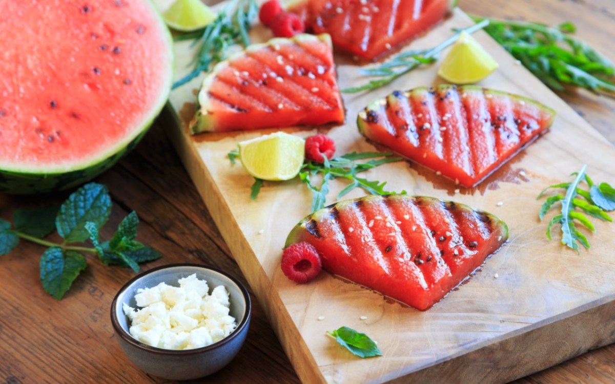 Wassermelonenstücke vom Grill auf einem Holzbrett. Daneben steht eine Schale mit Feta.