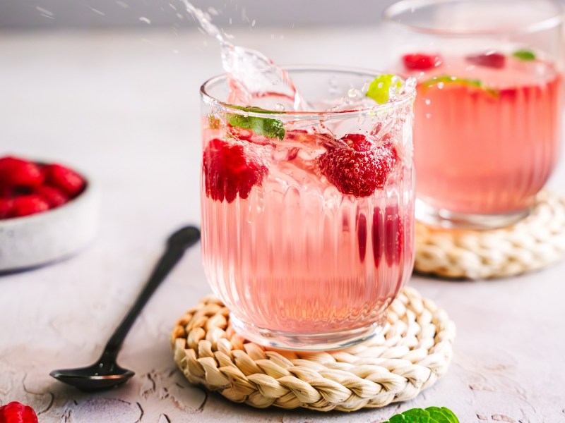 2 Gläser Wild Berry Spritz mit Himbeeren und Minze auf Untersetzern aus Korb, daneben ein Löffel.