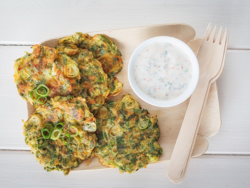 Zucchinipuffer auf einer Pappunterlage. Daneben ein Dip und eine Holzgabel.