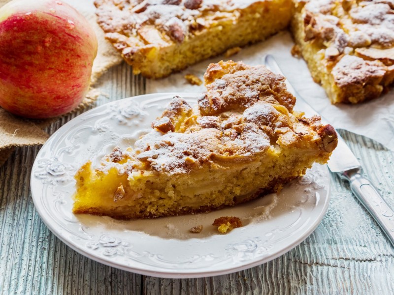 Ein Stück Apfelkuchen ohne Mehl auf einem Teller, daneben der angeschnittene Kuchen und ein Apfel sowie Besteck.