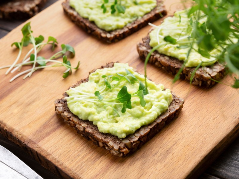 3 Scheiben Schwarzbrot mit Avocado-Dip mit Schmand bestrichen, darauf Kresse, die Brote liegen auf einem Brett.