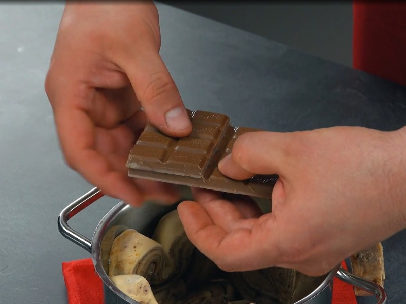 Blätterteigkuchen mit Schokolade aus dem Topf: Teigstücke in einem Topf, über dem zwei Hände eine Tafel Schokolade zerbrechen