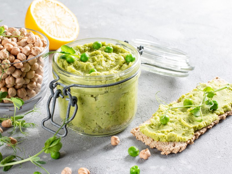 Erbsen-Hummus im Glas garniert mit Kresse, daneben Kichererbsen im Glas, eine halbe Zitrone und ein Knäckebrot mit Hummus bestrichen.