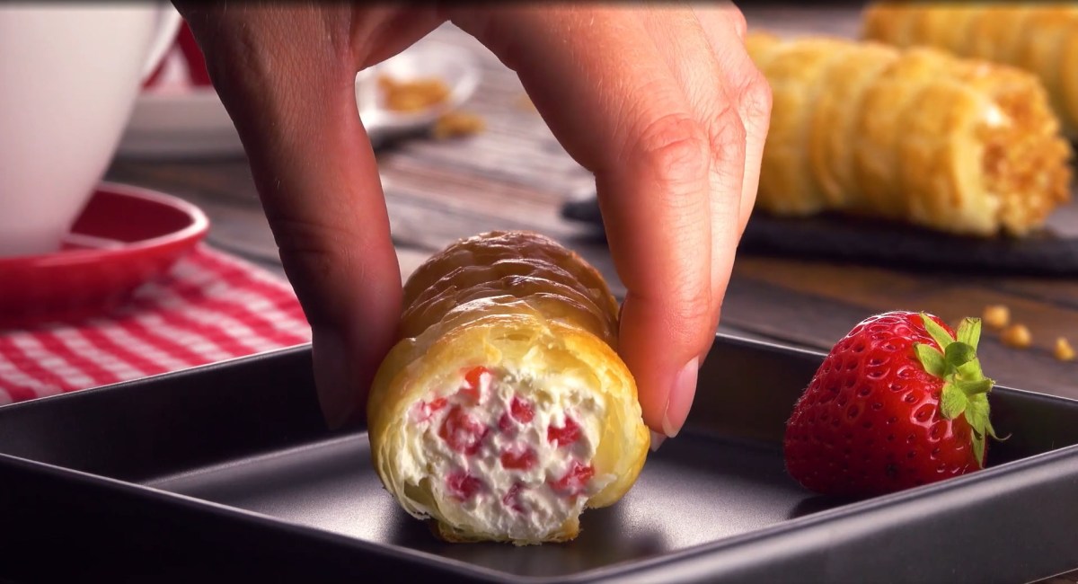 Ein Erdbeer-Röllchen wird auf einen dunklen Dessertteller gelegt, daneben eine Erdbeere.