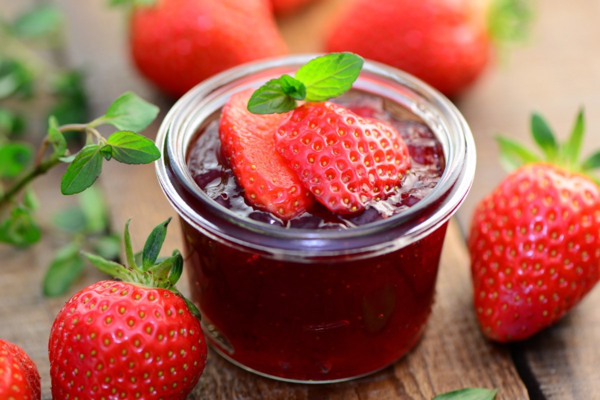 Ein Glas Erdbeermarmelade mit Minze und frischen Erdbeeren on top. Drumherum liegen Erdbeeren.