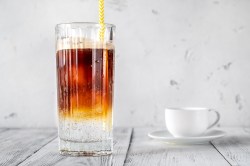 Ein Glas Espresso Tonic mit Eis und Strohhalm, daneben eine weiße Tasse mit Untertasse.