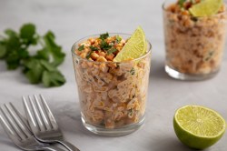 2 Gläser Esquites, mexikanischer Maissalat, garniert mit Limettenscheiben und Koriander, daneben zwei Gabeln, frischer Koriander und eine halbierte Limette.