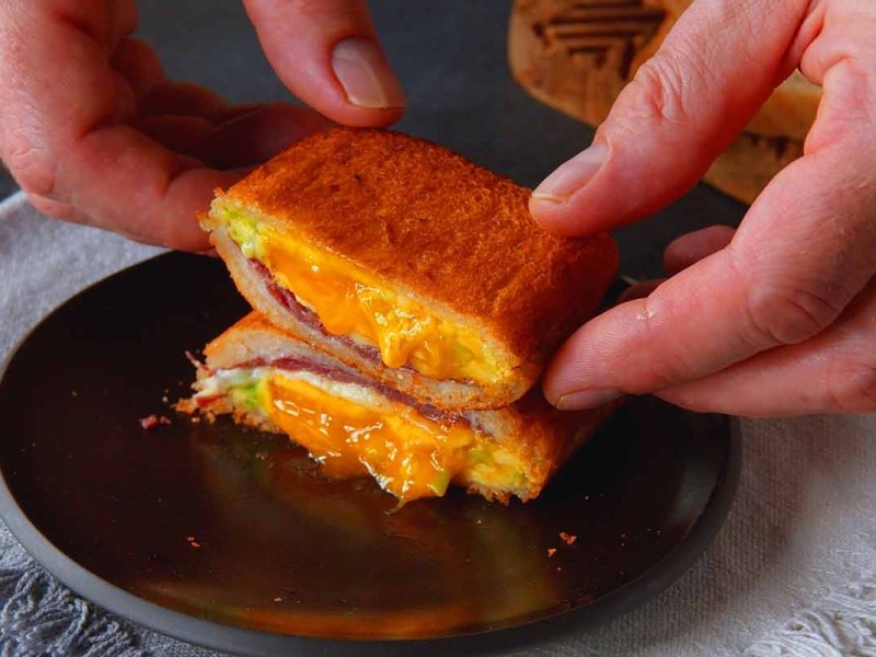 Ein gebackenes Sandwich auf einem Teller, das halbiert wurde und von zwei Händen gehalten wird.