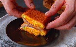 Ein gebackenes Sandwich auf einem Teller, das halbiert wurde und von zwei Händen gehalten wird.