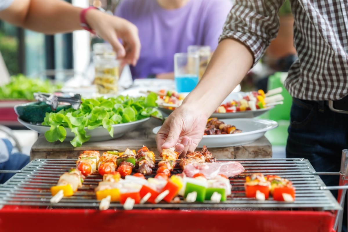 Was zu einem Grillabend dazu gehört: ein Grill mit Spießen im Vordergrund. Dahinter Beialgen und Menschen.