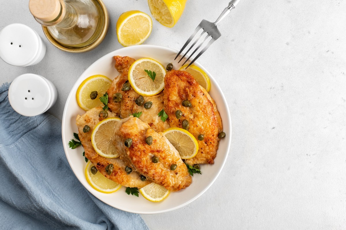 Piccata mit HÃ¤hnchen auf einem weiÃŸen Teler. Daneben liegt Zitrone und steht eine Flasche OlivenÃ¶l.